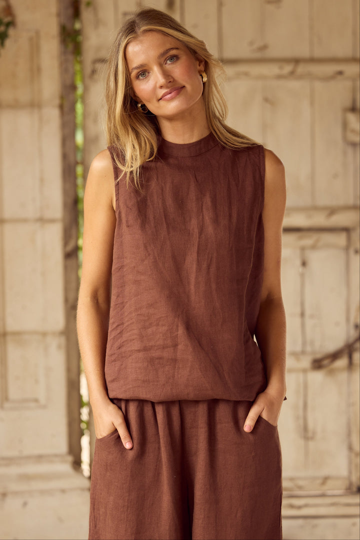 A woman wearing a sleeveless rust-brown linen top and matching wide-leg linen pants, standing in front of a rustic light-colored wooden door.