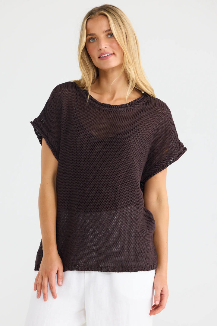A woman modeling a dark brown short-sleeve knit top with a sheer mesh overlay and white pants, photographed against a plain white studio background.