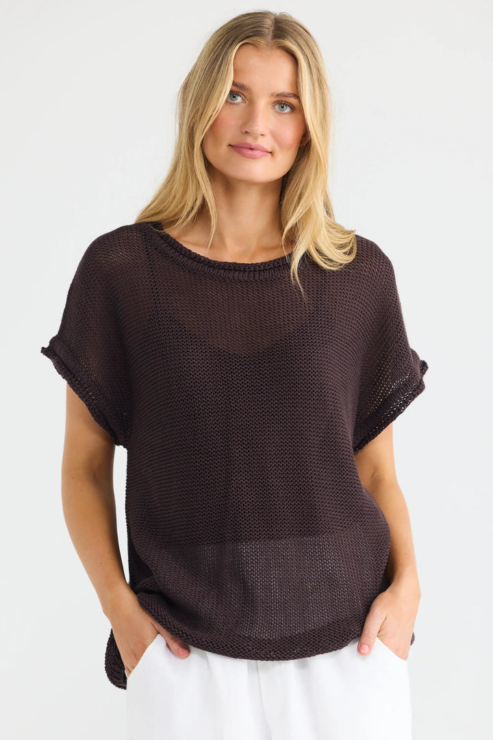 A woman modeling a dark brown short-sleeve knit top with white pants against a plain white studio background.