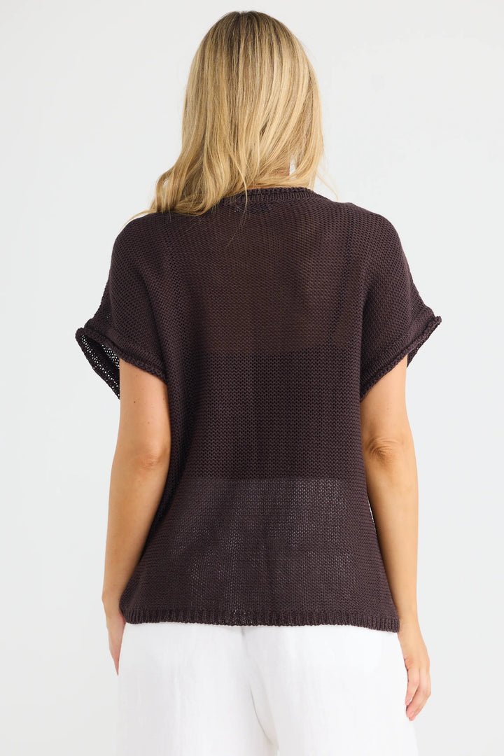 A woman viewed from the back wearing a dark brown short-sleeve knit top and white pants, standing against a plain light gray studio background.
