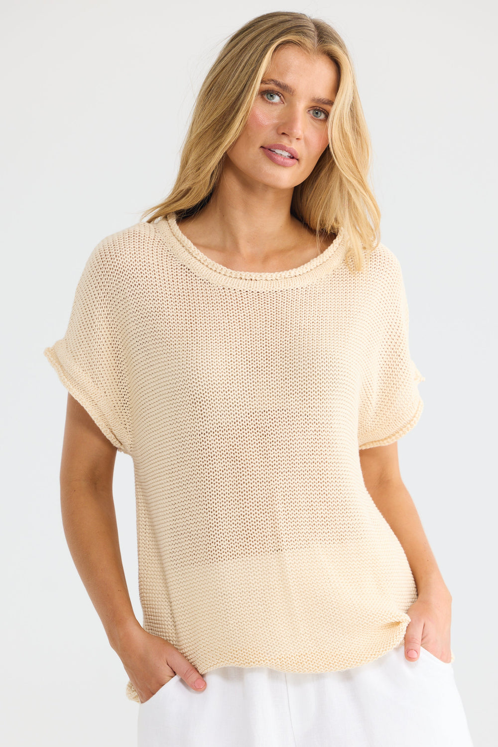 A woman modeling a short-sleeve, open-knit beige top with white pants against a plain white studio background.