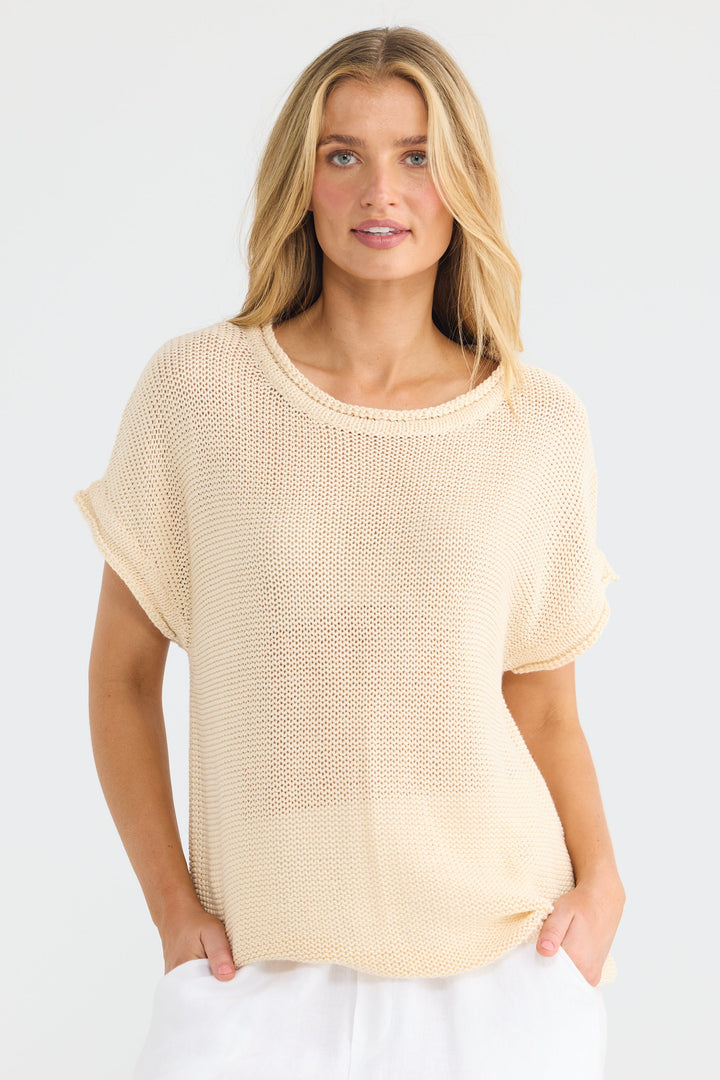 A woman modeling a short-sleeve, open-knit crewneck top in a light cream or beige color, paired with white pants, against a plain white studio background.
