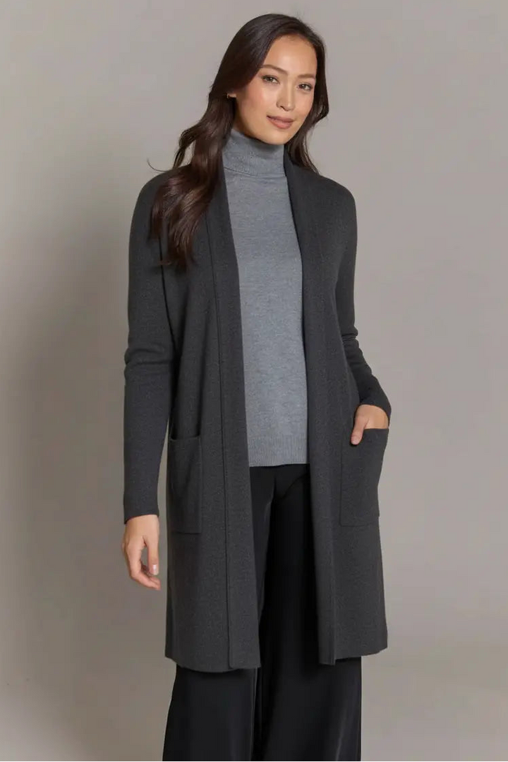 Woman wearing a long dark gray coat over a gray turtleneck and black pants on a plain background