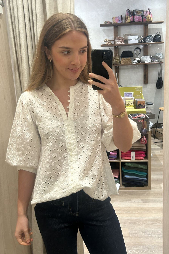 Woman taking a mirror selfie wearing a white blouse with black pants in a store.