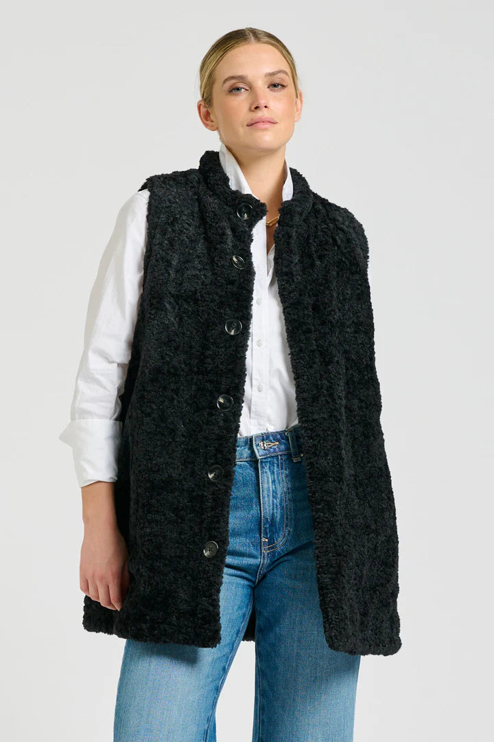 Woman wearing a black textured vest over a white shirt and blue jeans on a light gray background