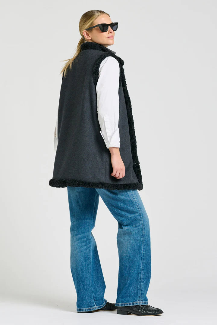 Person wearing a black vest with fur trim, white shirt, blue jeans, and sunglasses on a white background
