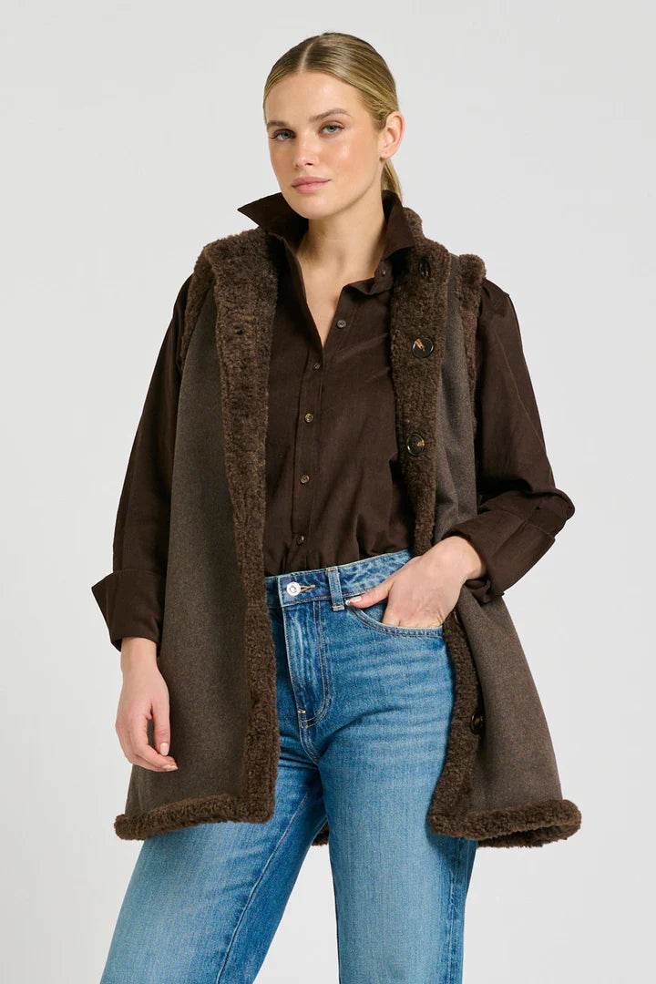 Woman wearing a brown coat with fur lining, brown shirt, and blue jeans on a gray background