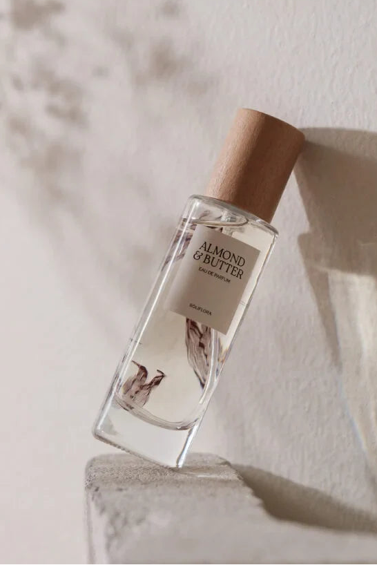 A clear rectangular glass perfume bottle labeled 'ALMOND BUTTER' with a light wood cap, leaning against a textured off-white wall with soft shadows.