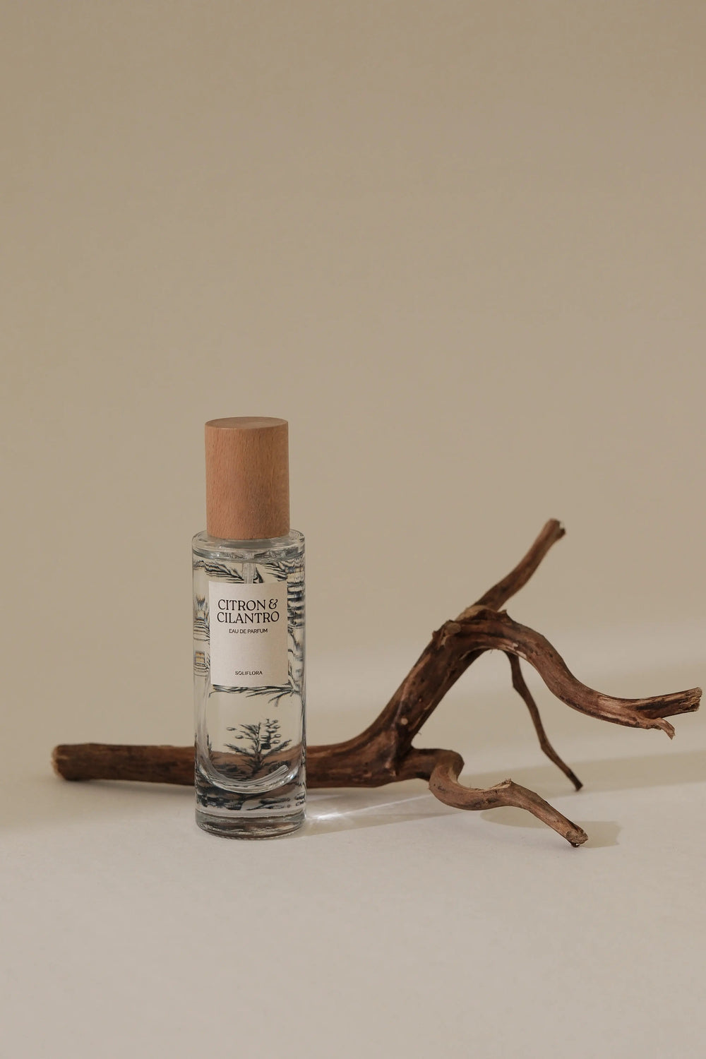 A clear glass roll-on bottle with a natural cork cap and a label reading 'Citronel Odorifer' and 'CITRONEL OLIOUS', styled on a beige surface with a piece of driftwood against a solid beige background.