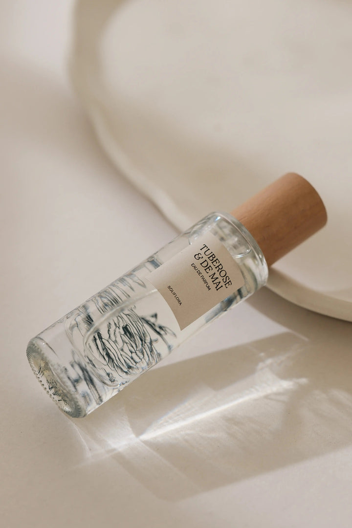 A clear glass perfume bottle with a light wood cap and a cream label, lying diagonally on a warm beige surface with a soft white ceramic dish in the background.