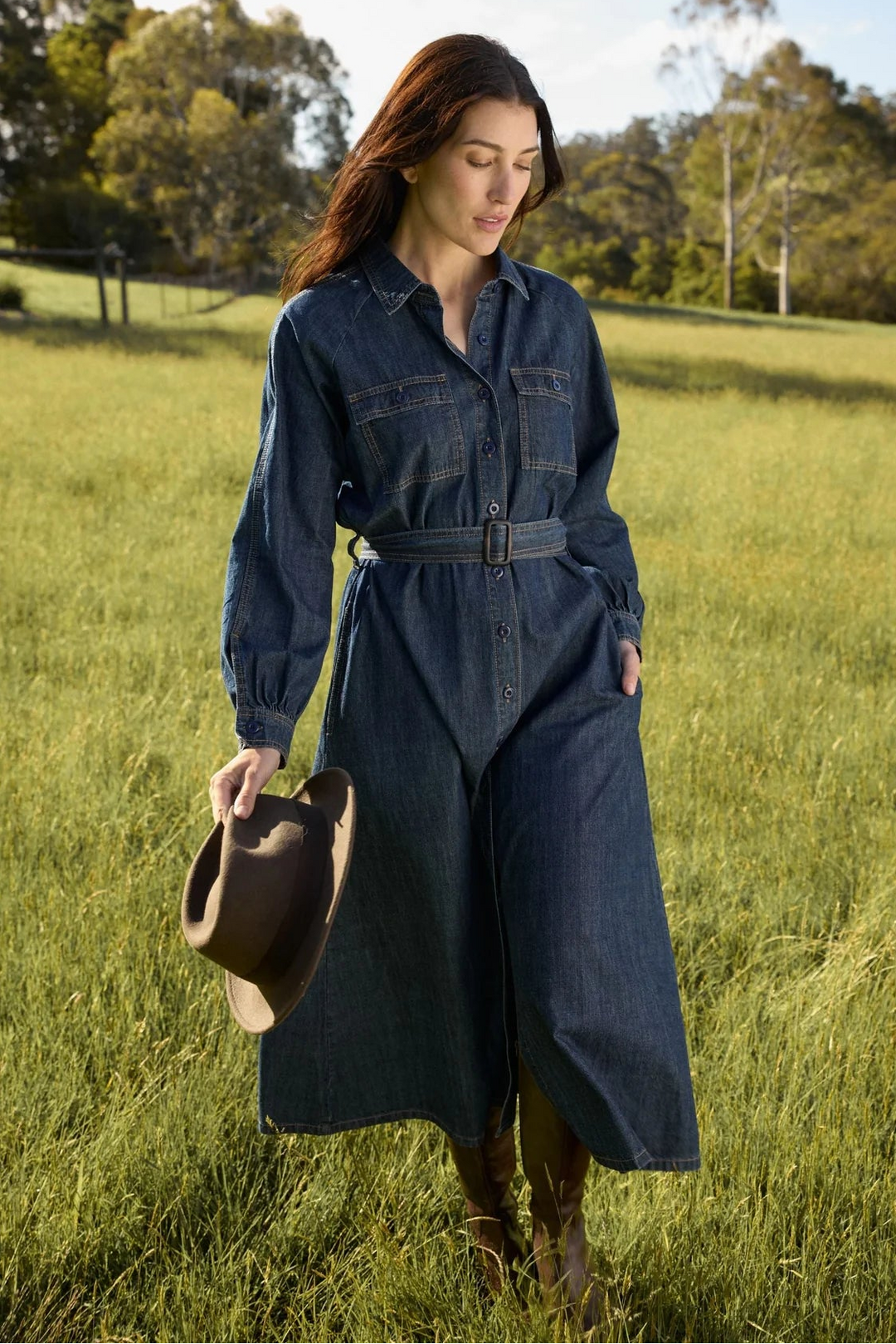 Woman in a denim dress standing in a grassy field holding a hat.