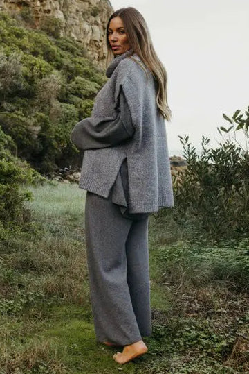 Person in a gray outfit standing in a natural setting with greenery and mountains.
