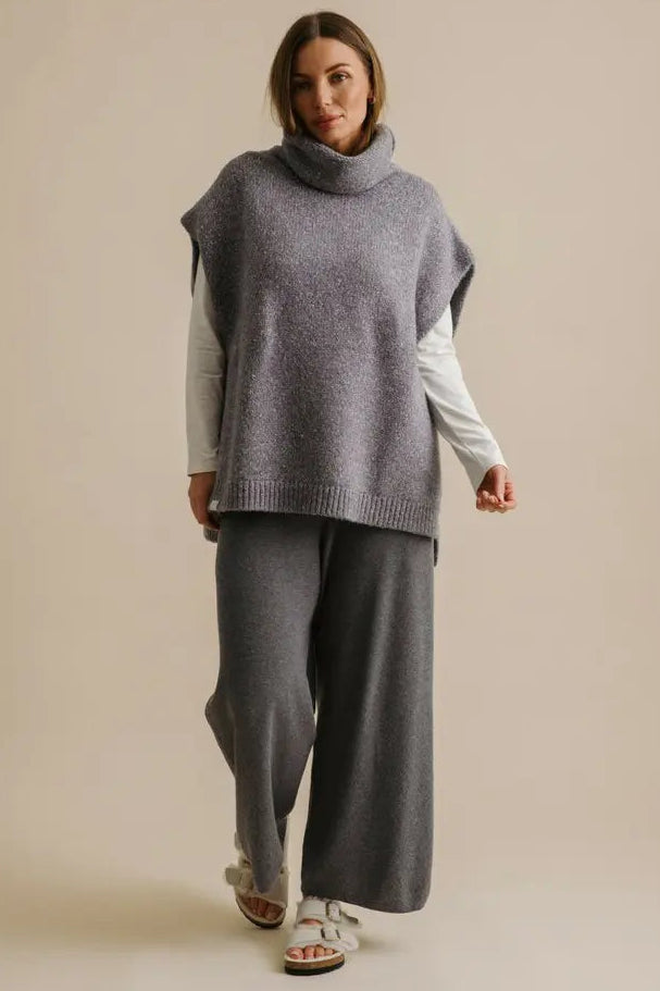 Woman wearing a gray sleeveless sweater over a white shirt with gray pants on a beige background