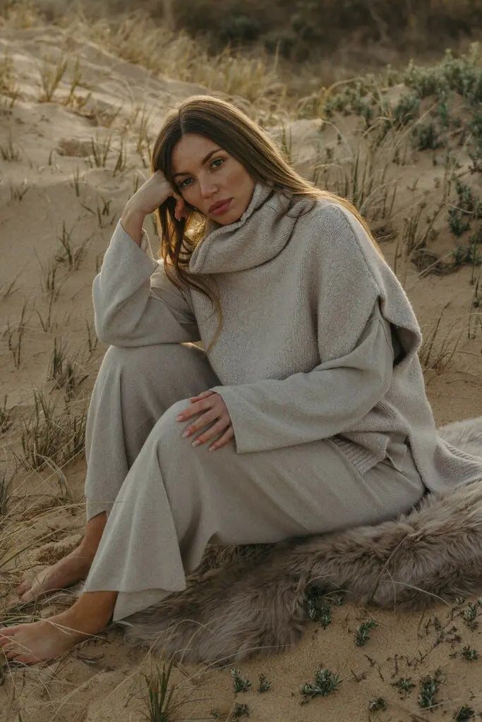 Woman in a beige outfit sitting on sand dunes during sunset.