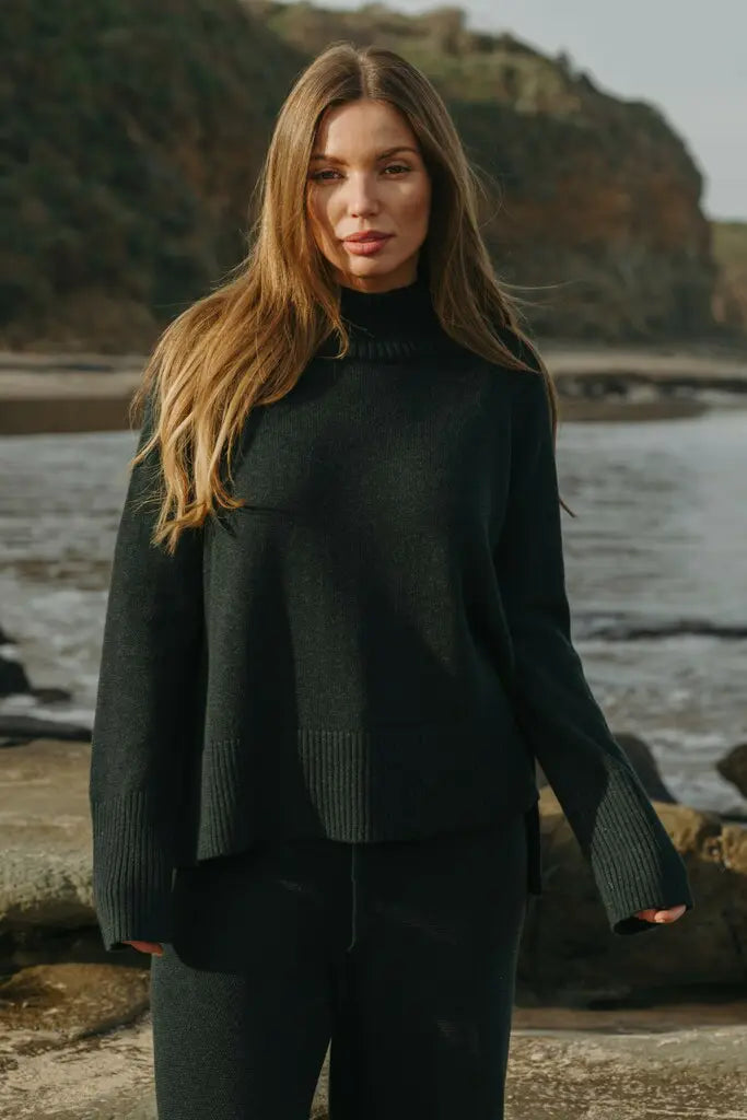 Woman wearing a dark green sweater standing by a body of water with cliffs in the background