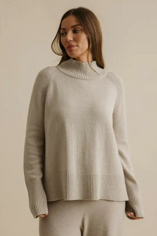 Woman wearing a beige sweater and pants against a plain background