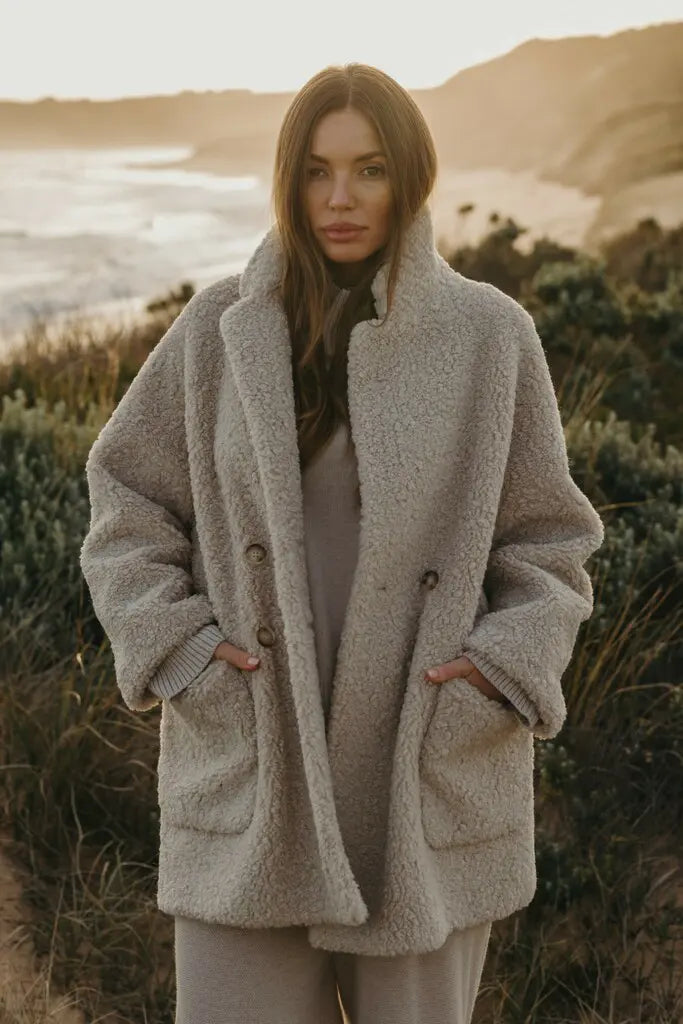 Woman wearing a beige coat standing in a natural setting with mountains and water in the background