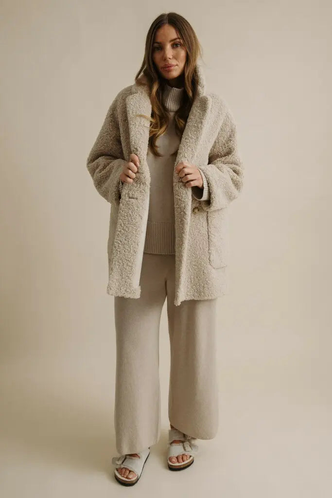 Woman wearing a beige coat, sweater, and pants against a plain background