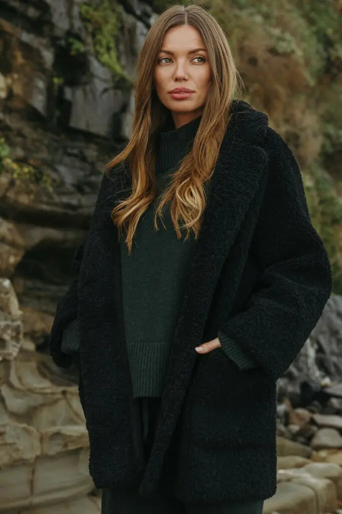 Woman wearing a dark coat standing in a natural setting with rocks and greenery.