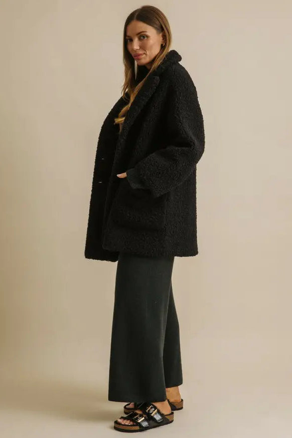 Woman wearing a black coat and dark pants against a beige background