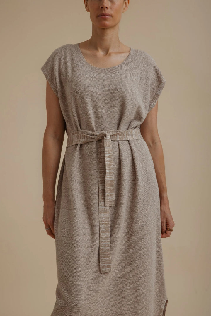 A woman wearing a light taupe linen-blend dress with a matching fabric tie belt at the waist, photographed against a solid beige studio background.