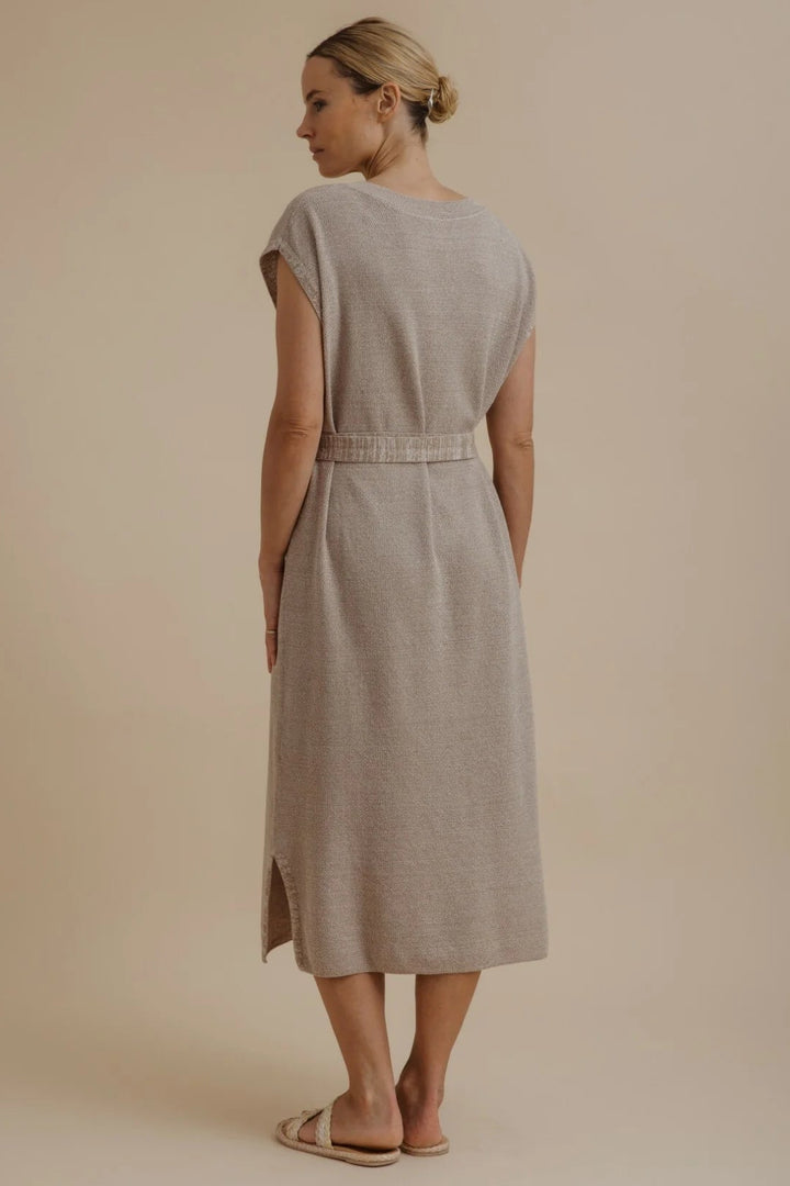 A woman wearing a light beige linen midi dress with a tie waist, shown from the back against a solid beige studio background.