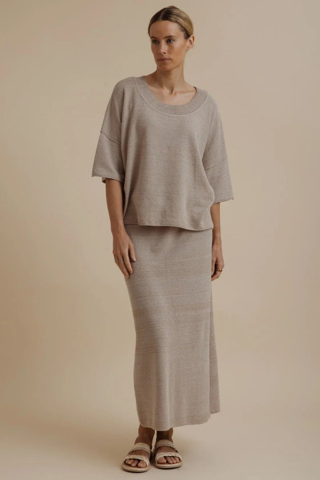 A woman modeling a matching two-piece linen set consisting of a short-sleeve cropped top and a long maxi skirt in a light taupe-beige color, set against a solid beige studio background.