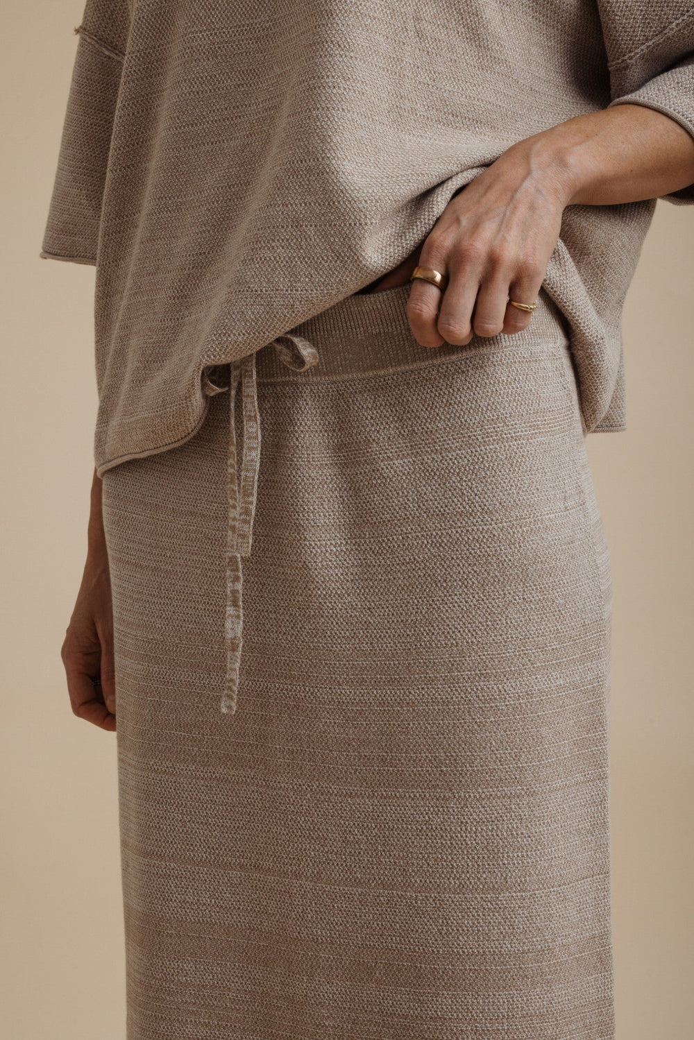 Close-up of a person wearing a textured taupe knit top and matching skirt with a drawstring waist, against a warm beige background.