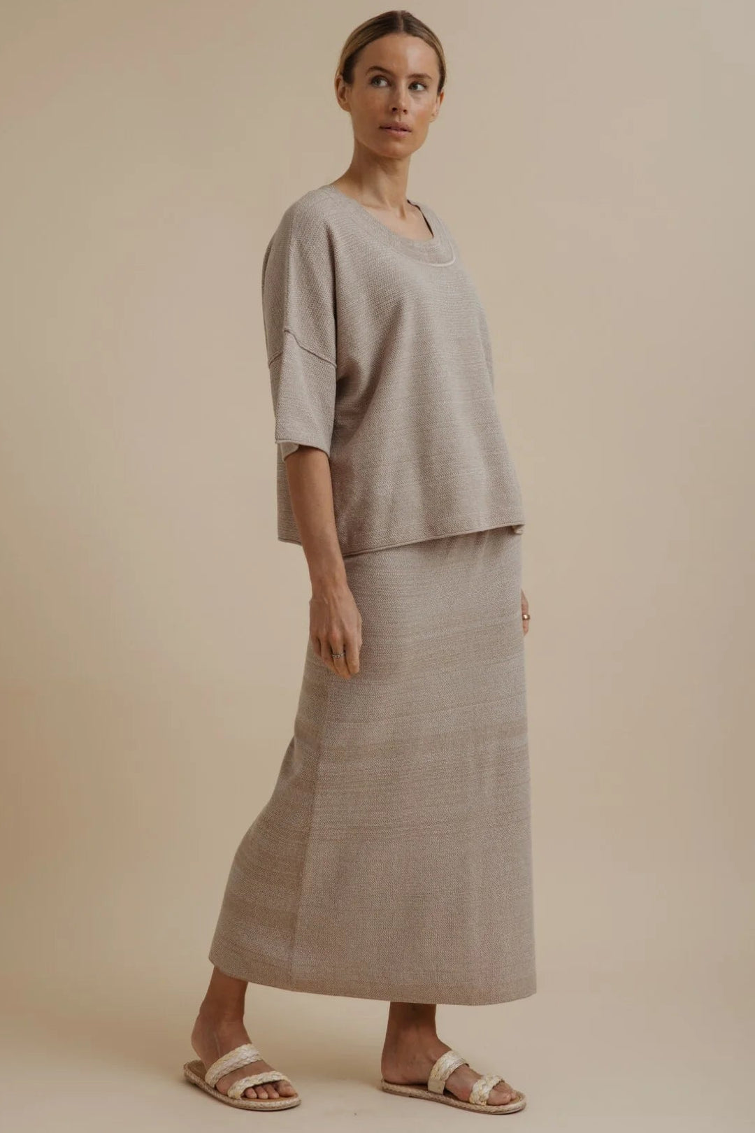A woman modeling a light beige linen two-piece set consisting of a short-sleeve top and a matching maxi skirt, paired with neutral sandals, against a solid beige studio background.