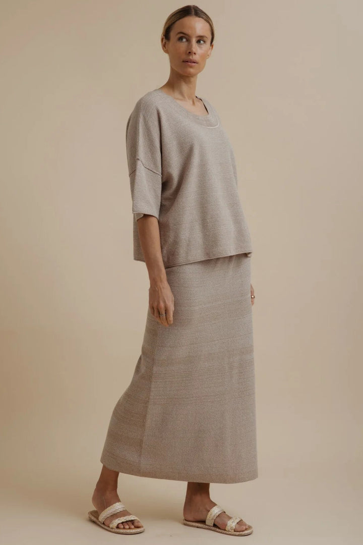A woman modeling a light beige linen two-piece set consisting of a short-sleeve top and a matching maxi skirt, paired with neutral sandals, against a solid beige studio background.