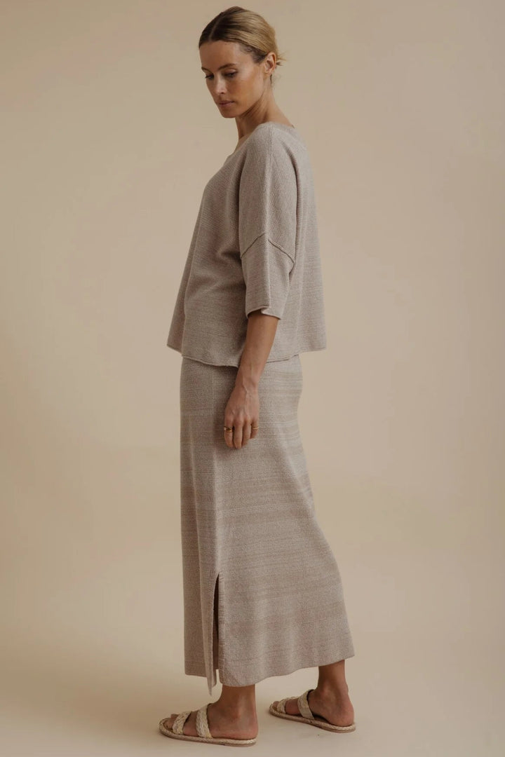 A woman modeling a light beige linen two-piece set consisting of a short-sleeve cropped top and a matching maxi skirt with a side slit, paired with neutral slide sandals, set against a solid beige studio background.