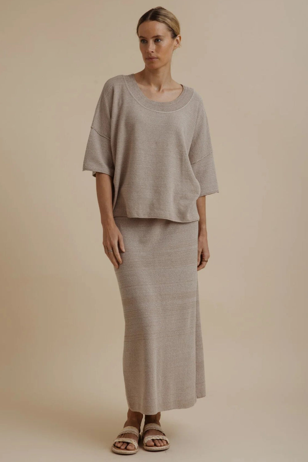 A woman modeling a matching two-piece linen set consisting of a short-sleeve cropped top and a long maxi skirt in a light taupe-beige color, set against a solid beige studio background.