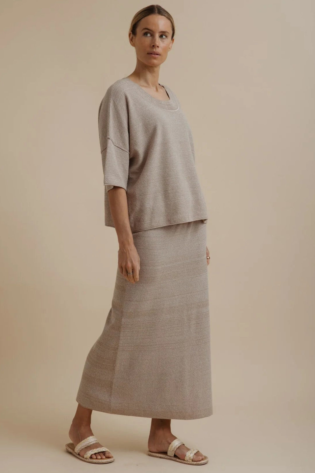 A woman modeling a light beige linen two-piece set consisting of a short-sleeve top and a matching maxi skirt, paired with neutral sandals, against a solid beige studio background.