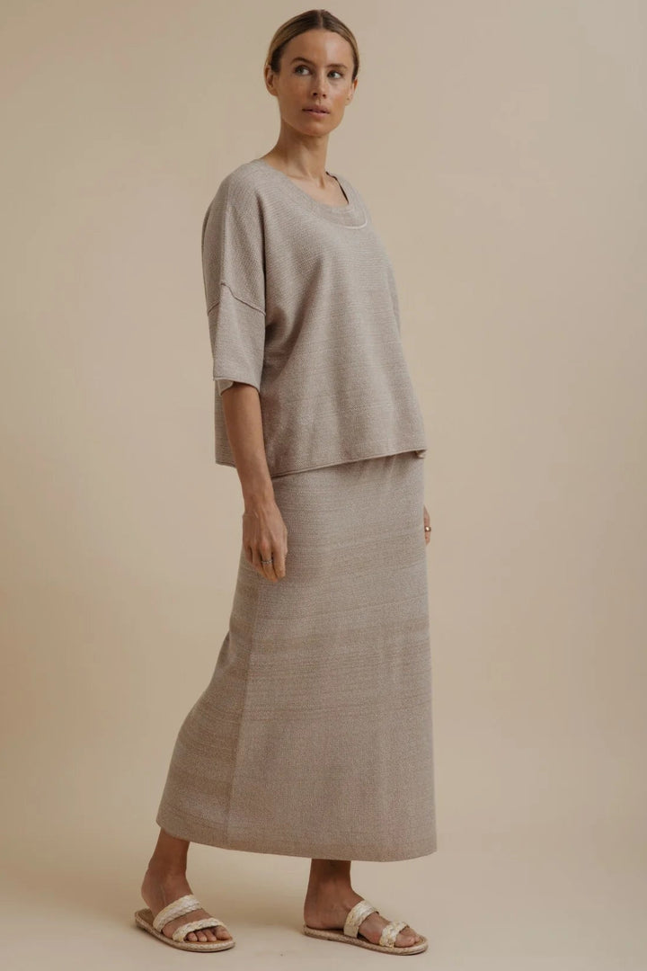A woman modeling a light beige linen two-piece set consisting of a short-sleeve top and a matching maxi skirt, paired with neutral sandals, against a solid beige studio background.