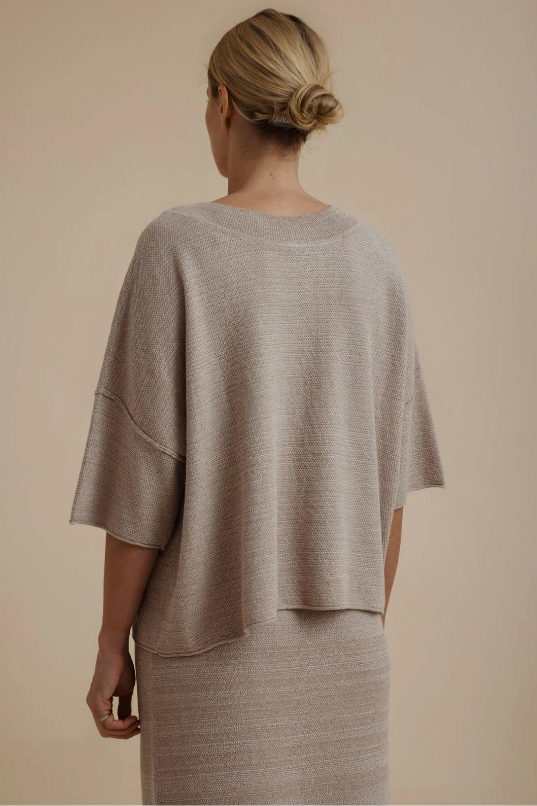 A woman shown from the back wearing a loose, short-sleeve knit top and matching textured skirt in a light taupe-beige color, set against a solid warm beige studio background.