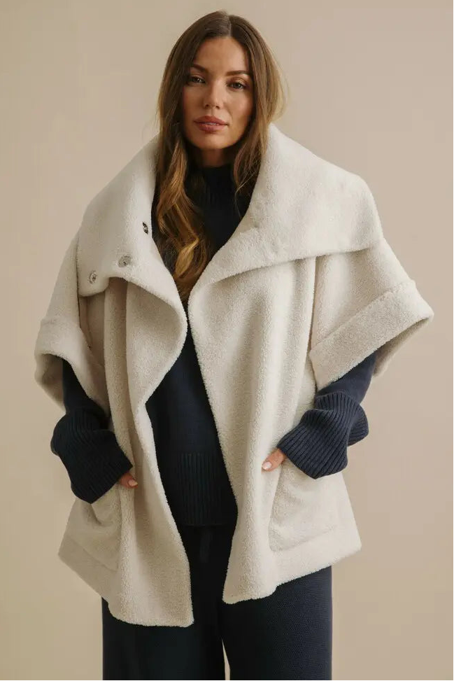 Woman wearing a cream-colored cape over a dark outfit against a beige background