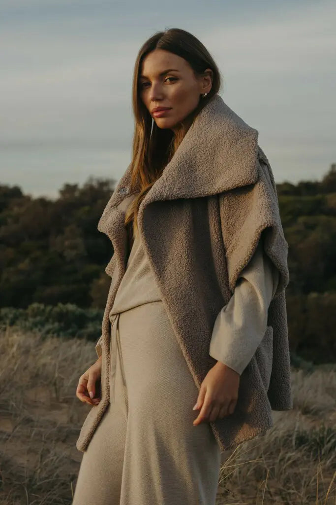 Woman wearing a beige coat standing in a natural setting with trees and sky.