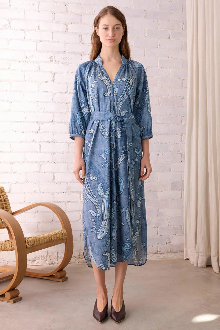 Woman wearing a blue patterned dress standing in a room with a white brick wall and wicker chair.