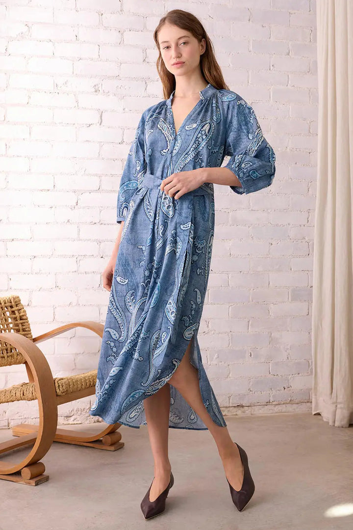 Woman wearing a blue patterned dress standing in a room with a white brick wall and wicker chair.