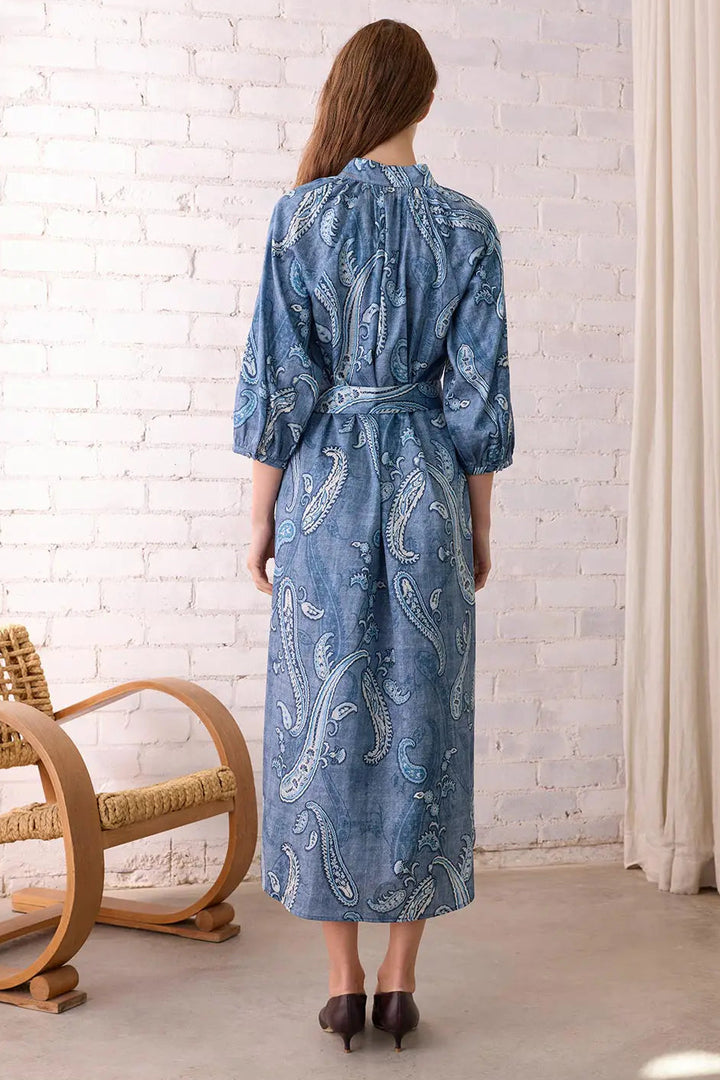 Woman wearing a blue patterned dress standing in a room with a white brick wall and wicker chair.