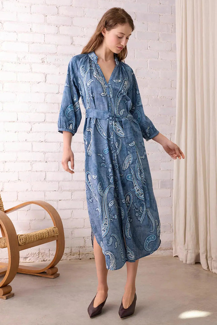 Woman wearing a blue patterned dress standing in a room with a white brick wall and wicker chair.