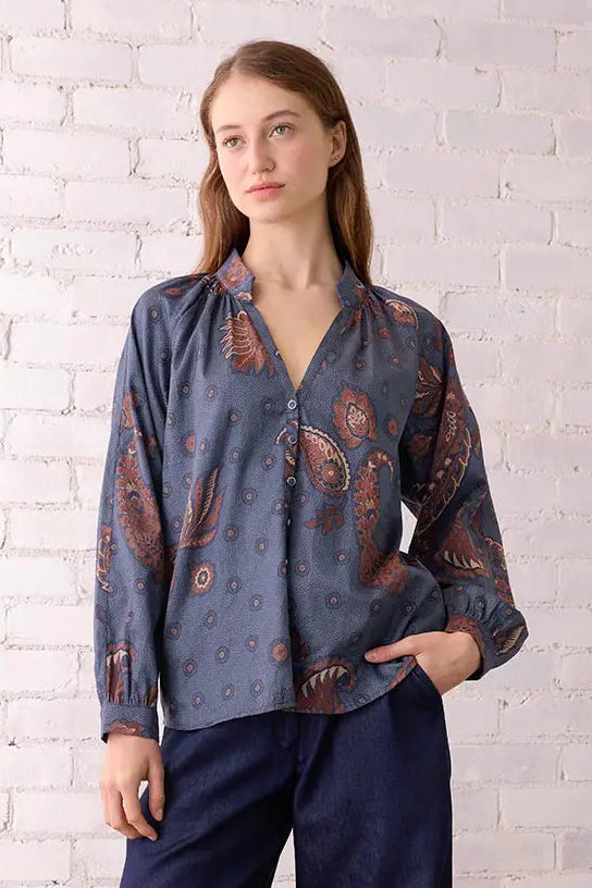 Woman wearing a patterned blouse and navy pants standing in a room with a white brick wall and wooden chair.