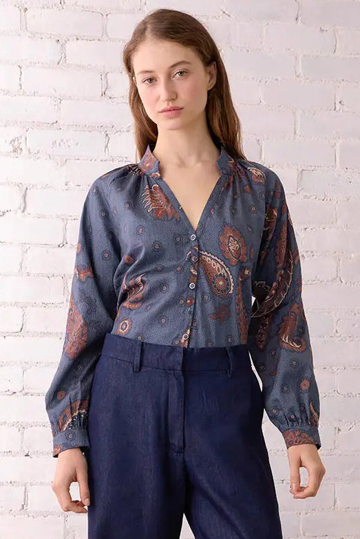 Woman wearing a patterned blouse and navy pants standing against a white brick wall.