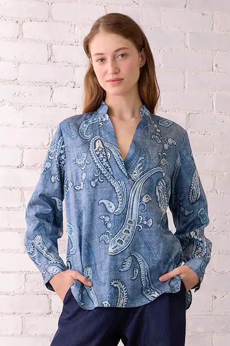 Woman wearing a blue embroidered blouse and dark pants in a room with a white brick wall and wooden chair.