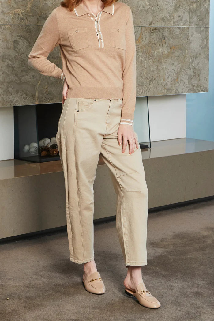 Woman wearing a beige sweater and pants standing against a stone wall.