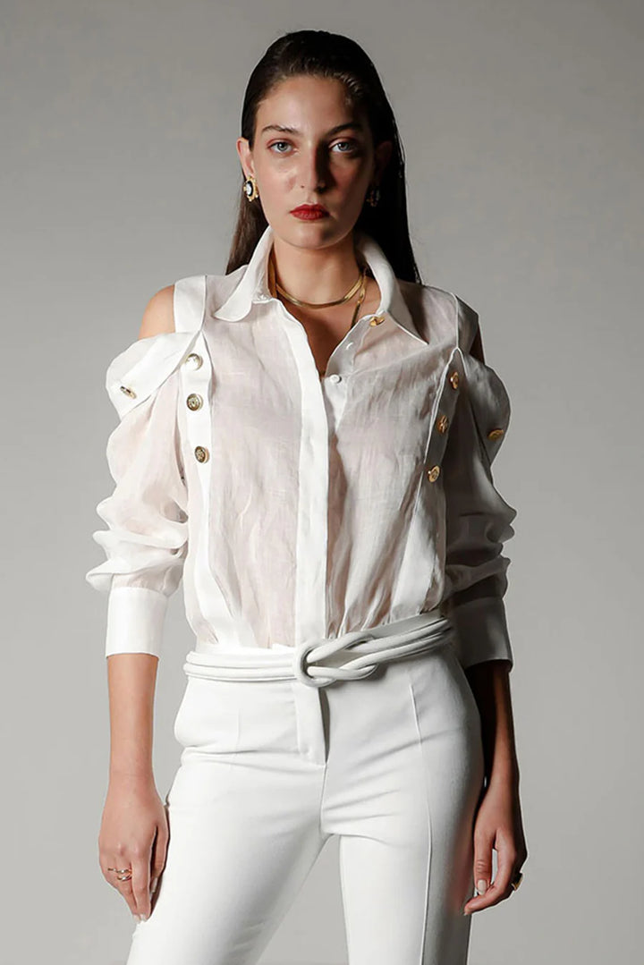 Woman wearing a white blouse with ruffled sleeves and a gray jacket over it, standing against a plain background.