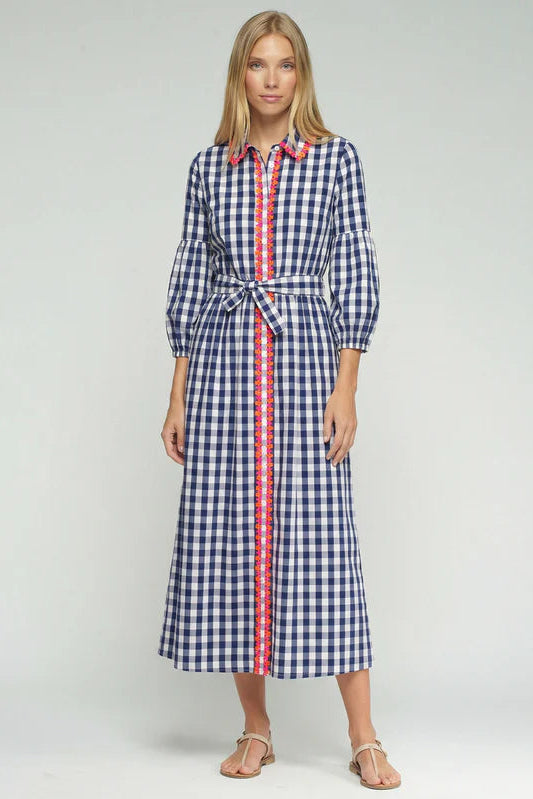 A woman modeling a long-sleeved midi shirt dress with a blue and white gingham pattern and a red and white striped center placket and tie belt, set against a plain light gray studio background.