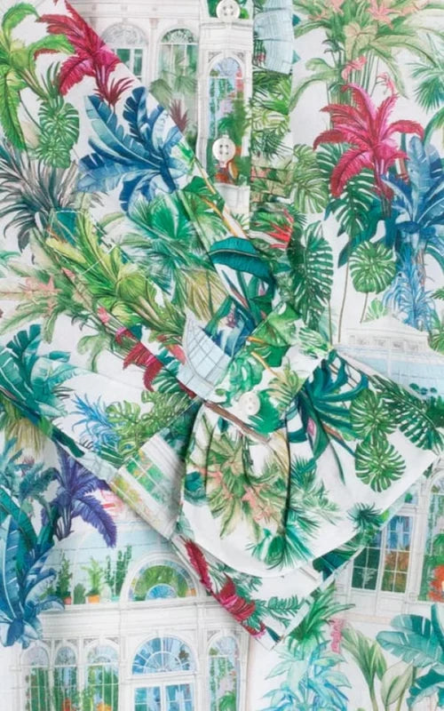 A close-up of a white fabric with a vibrant tropical print featuring green palm and monstera leaves, pink flowers, and a white conservatory building design.