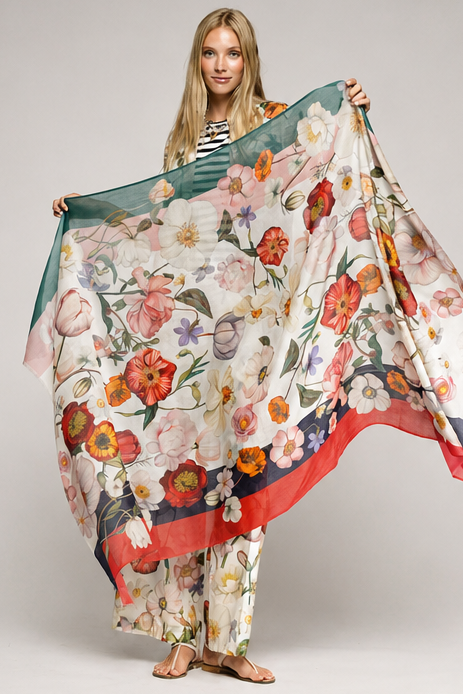 A woman modeling a large floral scarf with a matching printed pant set, holding the scarf wide open against a neutral gray studio background.