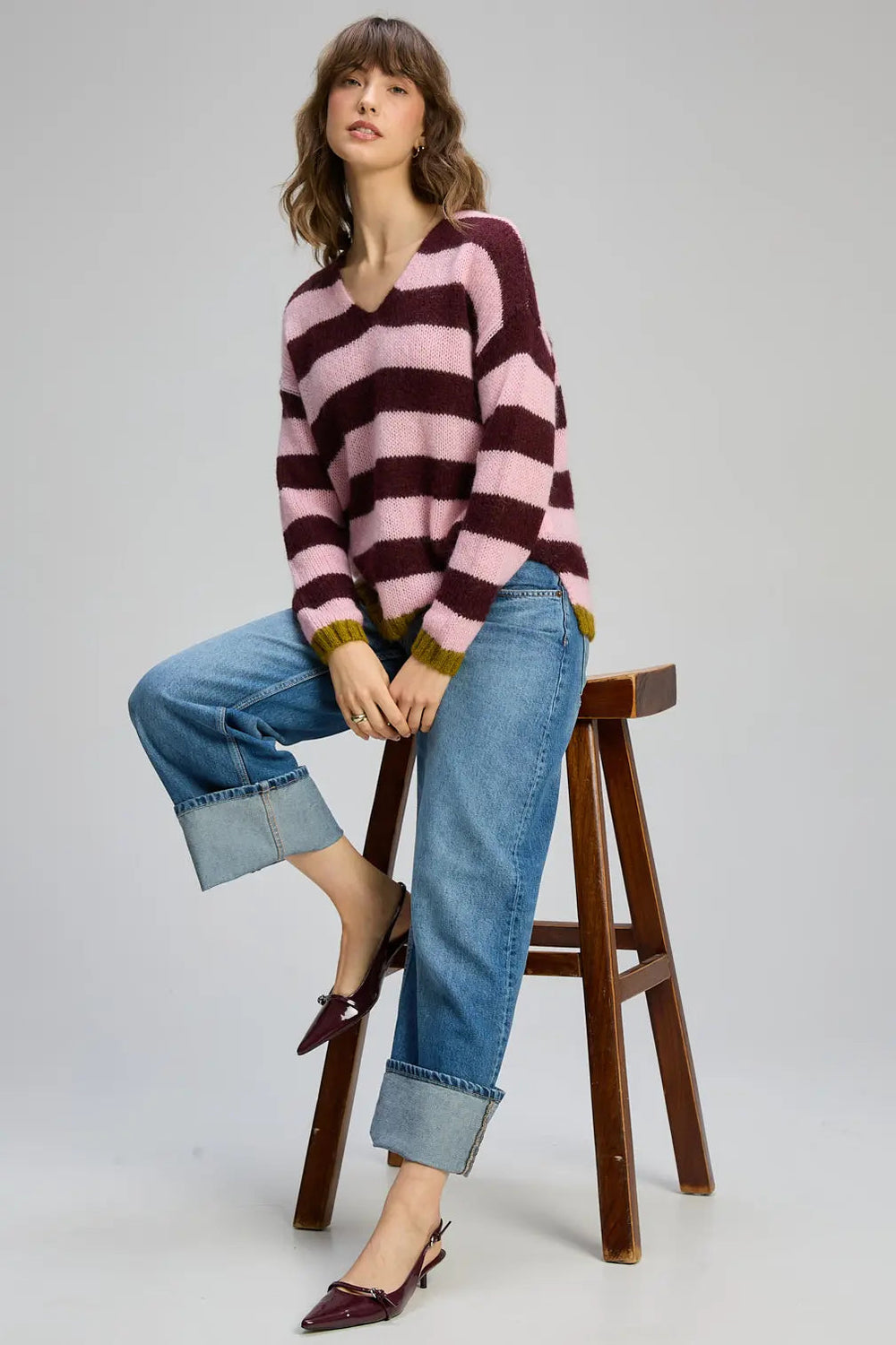 Woman wearing a striped sweater and jeans sitting on a wooden stool against a plain background